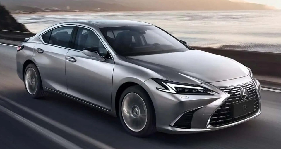 Silver Lexus ES, 2024 photo. The car is driving along a road by the water.